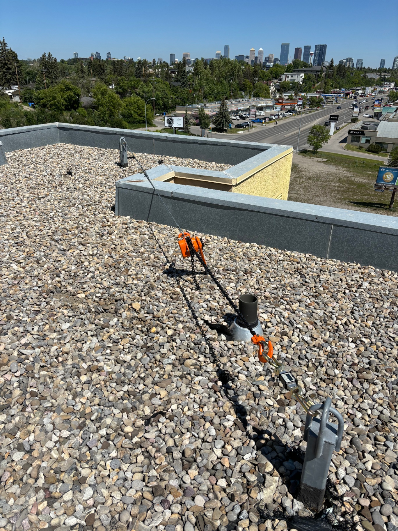 Chain-hoist pull test loading a single-point roof anchor with digital gauge in line, downtown Calgary skyline in the background