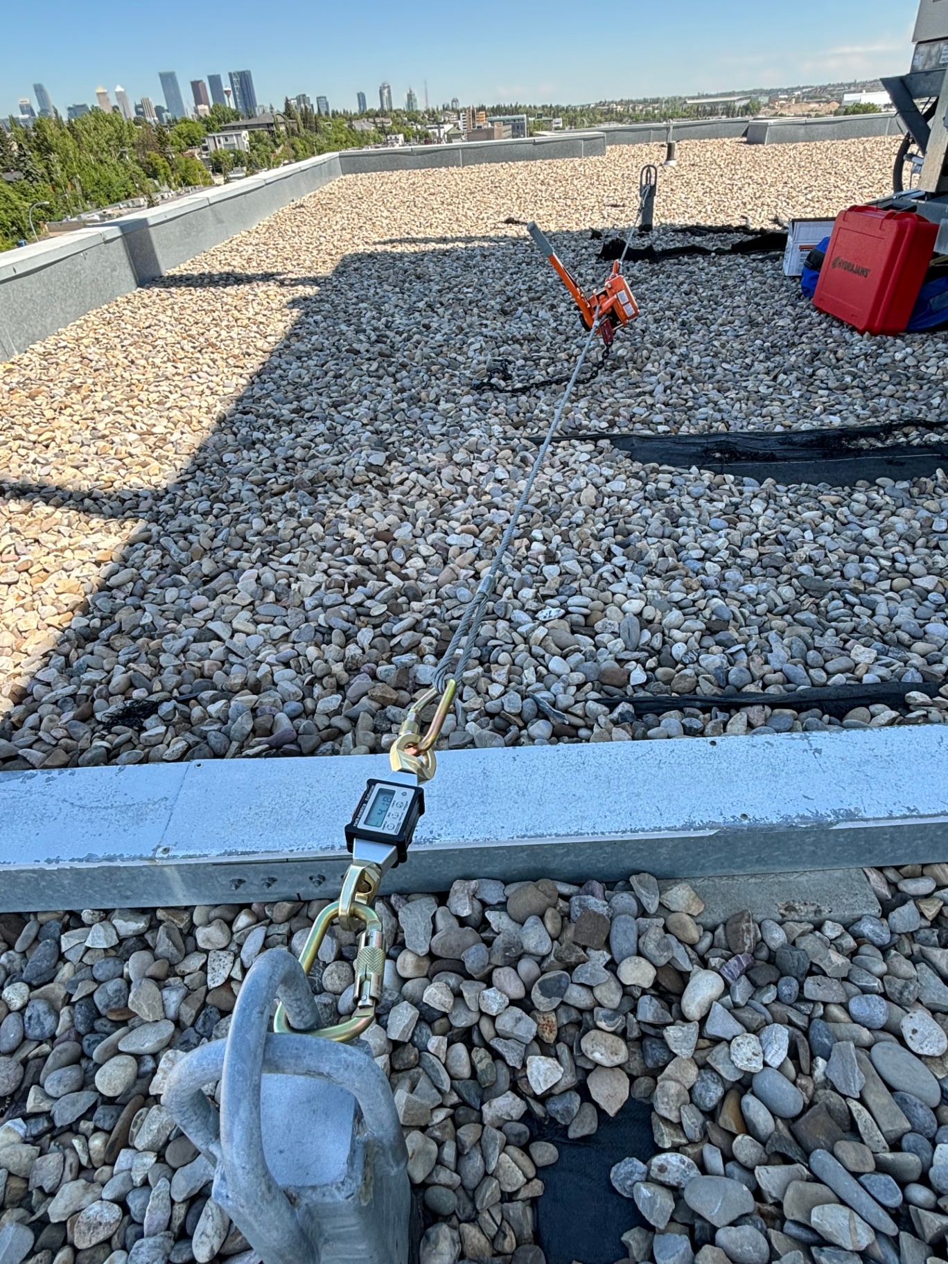 Hydrajaws cable pull-test rig anchored to a parapet beam with digital load gauge during single-point anchor proof loading on a Calgary rooftop