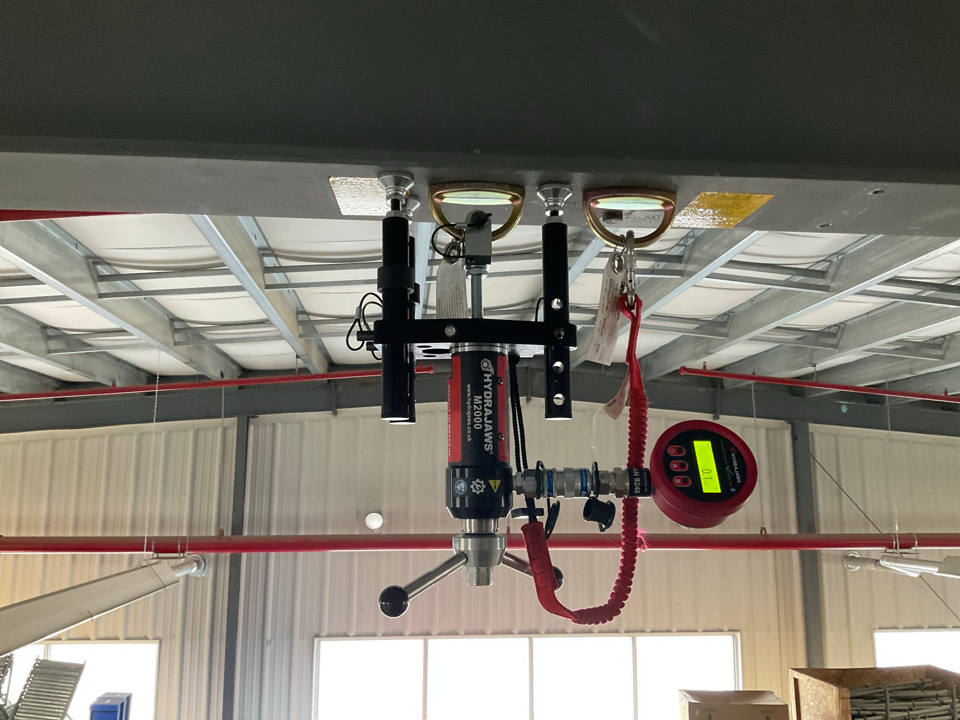 Hydrajaws M2000 pull-tester suspended from twin overhead ceiling anchors with red digital load gauge during proof loading inside a warehouse