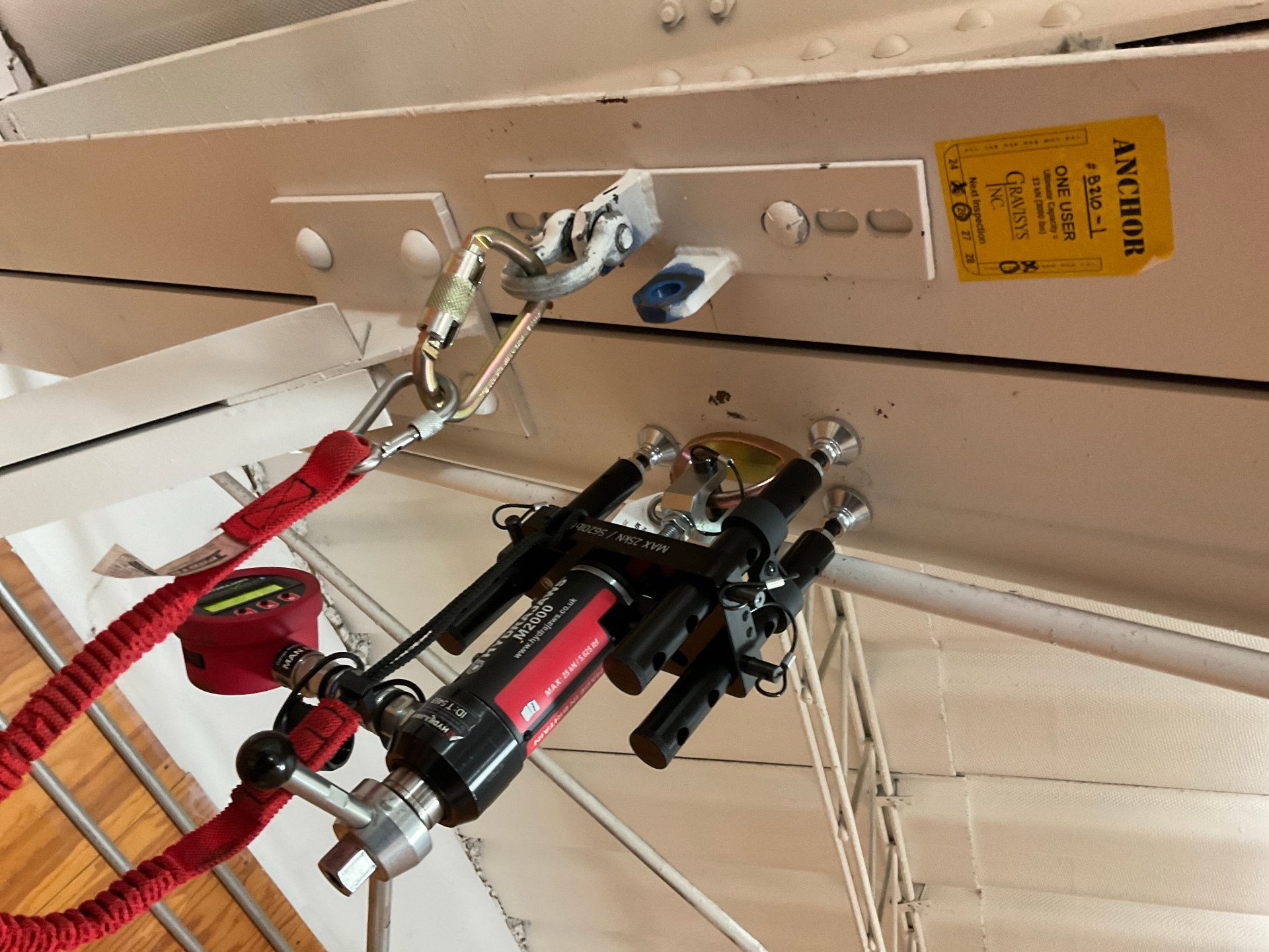 Close-up of an overhead beam-mounted anchor tag and Hydrajaws proof-loading rig connected via carabiner during single-point anchor pull testing